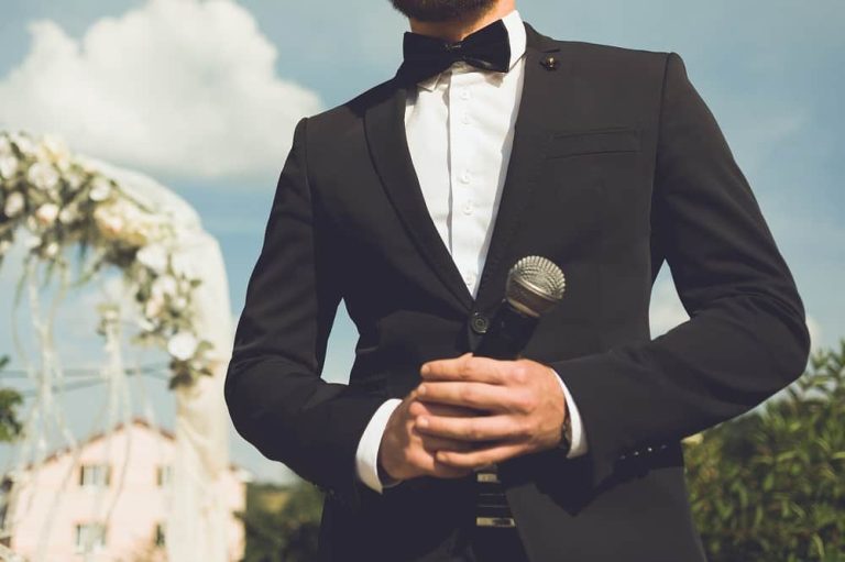 A man in a black suit and bow tie holds a microphone outdoors, with a wedding arch and house in the background, ready to introduce the Boston Band for unforgettable wedding music.