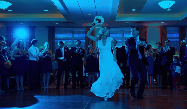A bride and groom dance in the center of a crowded reception hall, surrounded by applauding guests under blue lighting as lively wedding music from a Boston Band fills the air.
