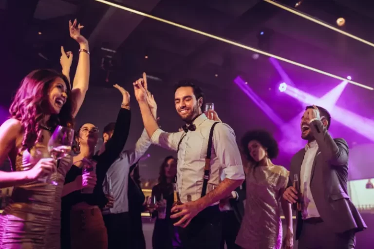 A group of people dressed in semi-formal attire dance, laugh, and hold drinks at a lively party with vibrant lighting, as a Boston Band delivers unforgettable wedding music.