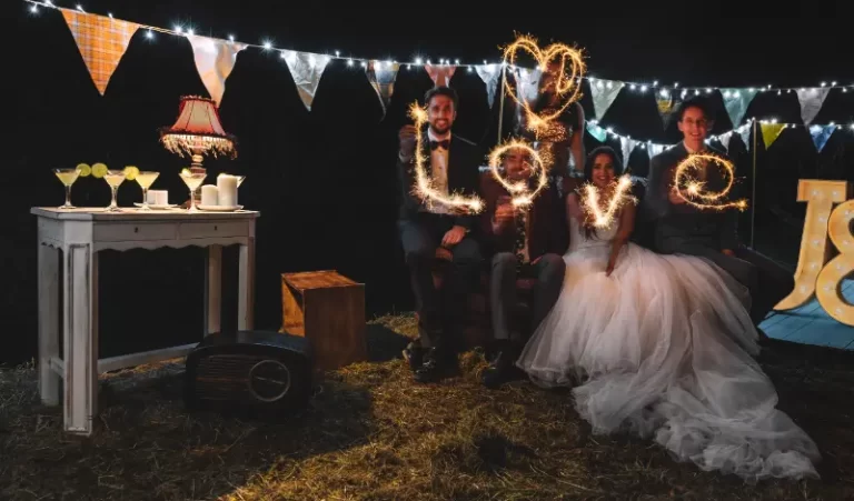 Four people, including a bride and groom, sit outdoors at night, holding sparklers that spell "love" in the air, as wedding music plays softly and string lights and decorations set the mood for this joyful Boston band celebration.