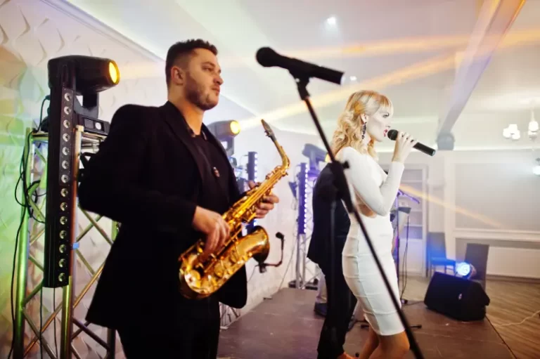 A man plays a saxophone while a woman in a white dress sings into a microphone on stage, with stage lights in the background—perfect entertainment from a talented Boston Band for unforgettable wedding music.