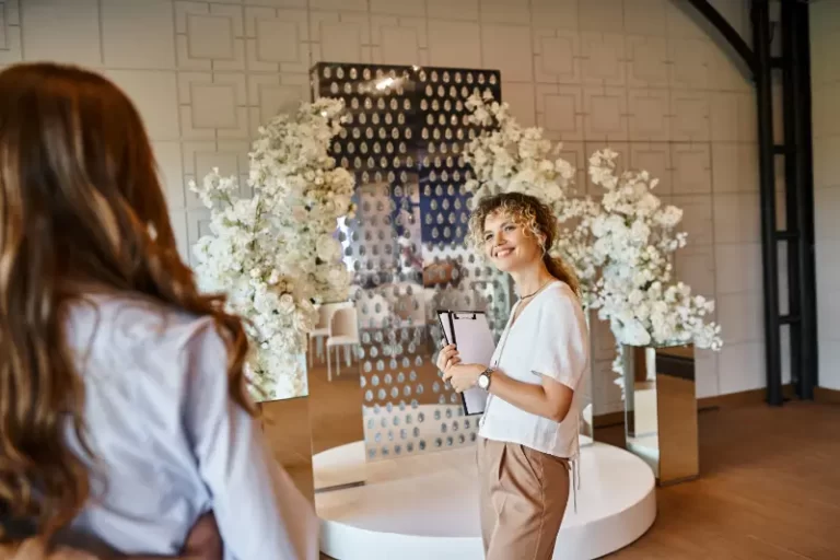 Two women stand near a decorative wedding arch with white flowers; one holds a clipboard and smiles while discussing the Boston Band playing the perfect wedding music for the event.