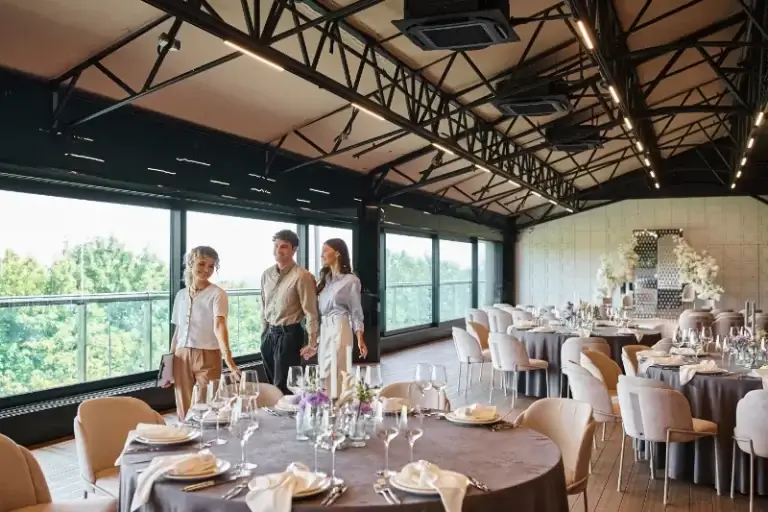 Three people walk into a decorated banquet hall with round tables set for an event, large windows on one side, floral arrangements by the wall, and the sound of wedding music from a Boston Band filling the air.