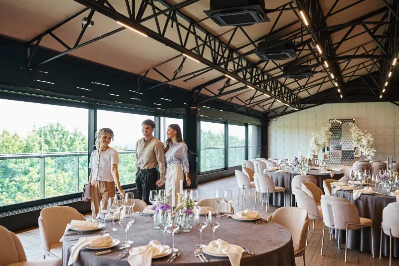 Three people walk into a decorated banquet hall with round tables set for an event, large windows on one side, floral arrangements by the wall, and the sound of wedding music from a Boston Band filling the air.
