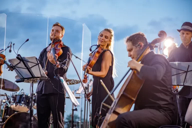 A Boston band of musicians performs outdoors, playing violins and a cello with music stands and equipment visible under evening sky lighting—perfect for elegant wedding music.