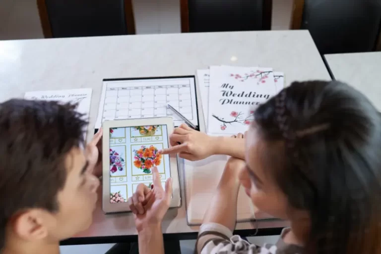 Two people sitting at a table review floral arrangements and discuss wedding music options on a tablet, with wedding planner documents and a calendar beside them.