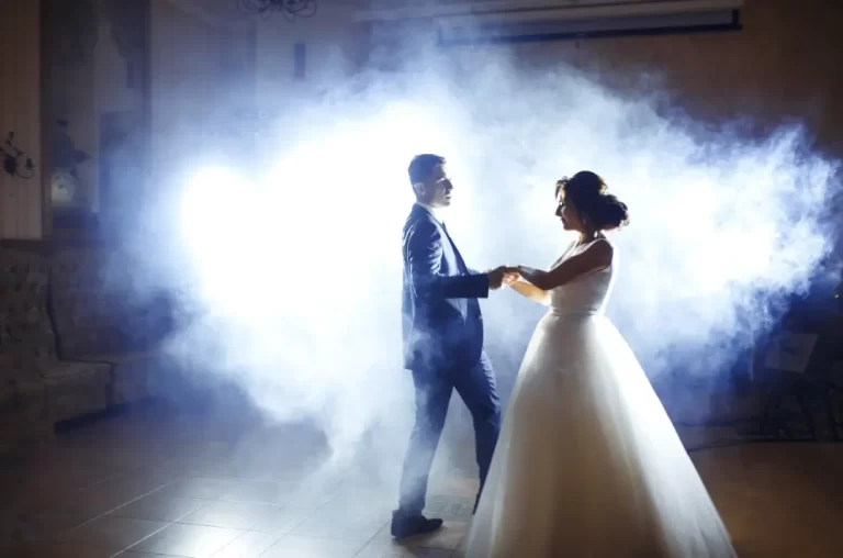 A bride and groom hold hands and dance on a dimly lit floor, surrounded by dramatic white fog and bright backlighting as a Boston Band fills the air with unforgettable wedding music.