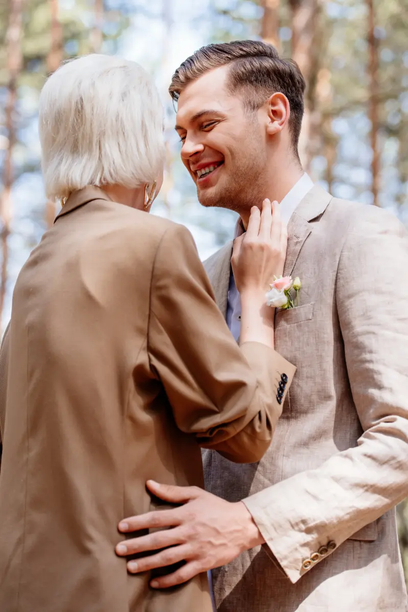 A couple dressed in beige suits stands close together outdoors, smiling at each other with trees in the background, as Wedding Music from a Boston Band drifts through the air.