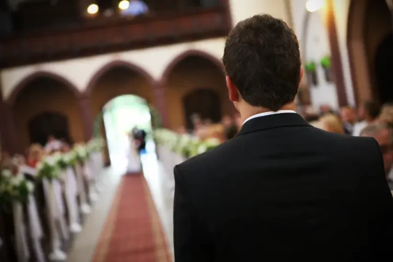 A man in a suit stands at the front of an aisle in a church, as a Boston Band softly plays wedding music while an approaching couple appears in the distance.
