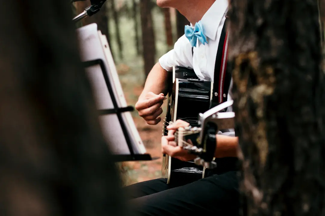 A person in a blue bow tie plays acoustic guitar outdoors, providing elegant wedding music with a music stand and trees visible in the foreground, perfect for any Boston Band performance.