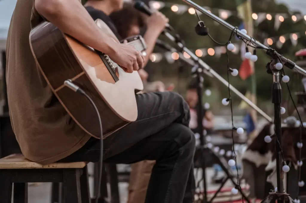 A person from a Boston Band plays acoustic guitar and sings into a microphone on an outdoor stage, string lights twinkling in the background—perfect for wedding music.
