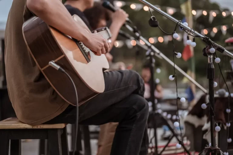 A person from a Boston Band plays acoustic guitar and sings into a microphone on an outdoor stage, string lights twinkling in the background—perfect for wedding music.