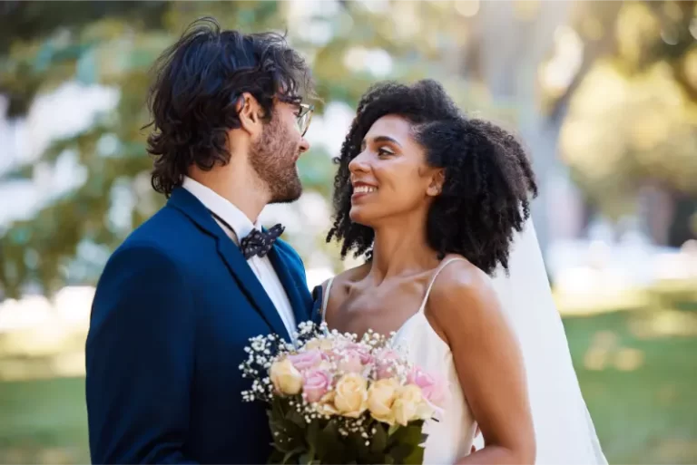 A bride and groom stand outdoors, smiling at each other as soft wedding music plays. The bride holds a bouquet of roses in her white dress and veil; the groom, in a blue suit and bow tie, looks blissful—celebrating their day with a top Boston band.