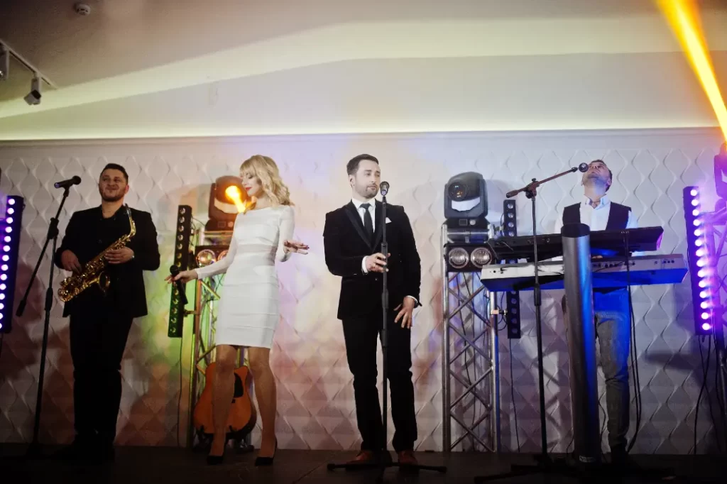Four musicians from a Boston band perform on stage; one plays saxophone, one sings, one stands at a microphone, and one plays keyboards. Stage lights and instruments in the background create an energetic wedding music vibe.