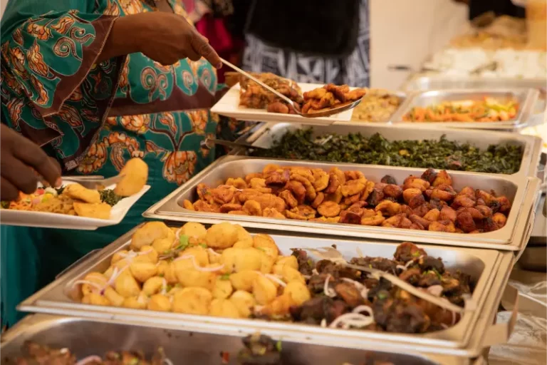 A person serves food from a buffet table filled with various dishes—including fried plantains, vegetables, and fried appetizers—while lively wedding music from a Boston band plays in the background.